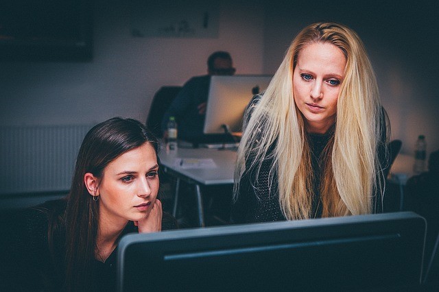 Two women working on a computer with a male in the background also working on a computer